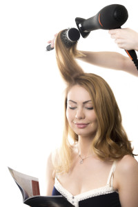 Portrait of young smiling gorgeous blond woman sitting at beauty salon reading fashion magazine, having her hair styled with hairdryer and round hairbrush, studio shot, isolated on white background