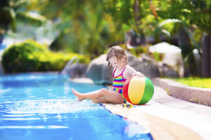 Adorable little girl with curly hair wearing a colorful swimming suit playing with water splashes at beautiful pool in a tropical resort having fun during family summer vacation